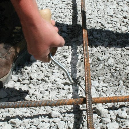 Close up action shot of a hand using the beige handle wire twister to secure a wire loop tie around intersecting rebar on a gravel base.