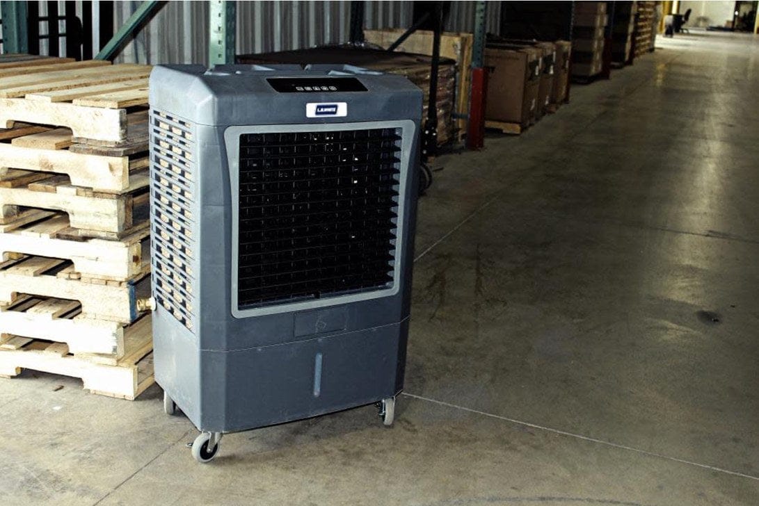 Large portable evaporative cooler in a warehouse setting with caster wheels, front-facing vent, and control panel, used for industrial cooling near shelving and pallets.