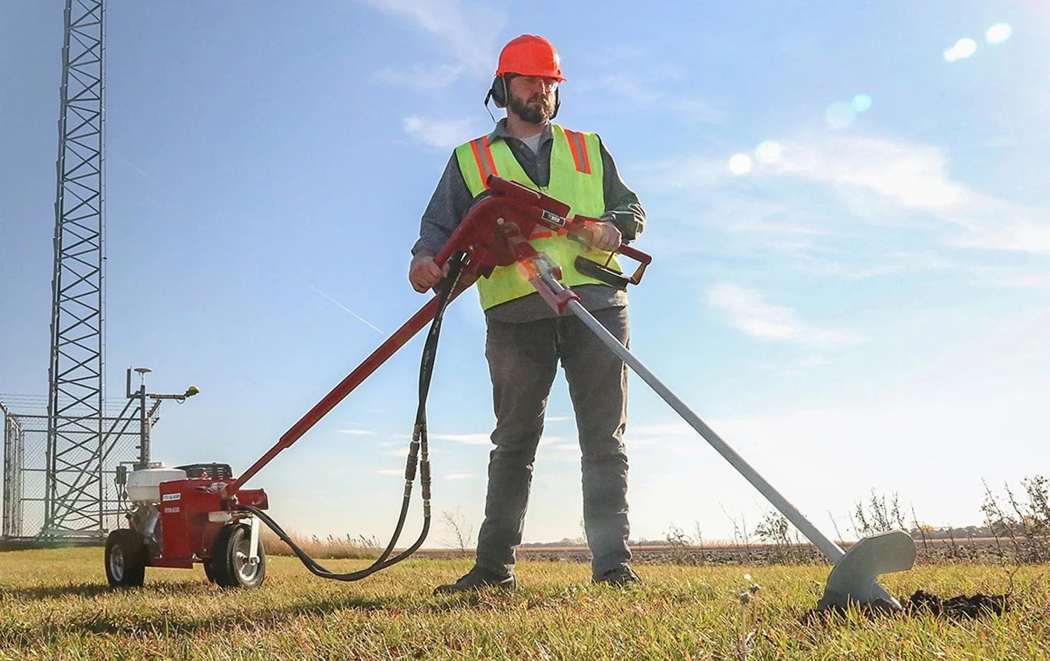 An operational demonstration showing a worker in safety gear using a Little Beaver hydraulic earth drill with a specialized attachment to drive a large ground anchor into a grassy field for structural support.