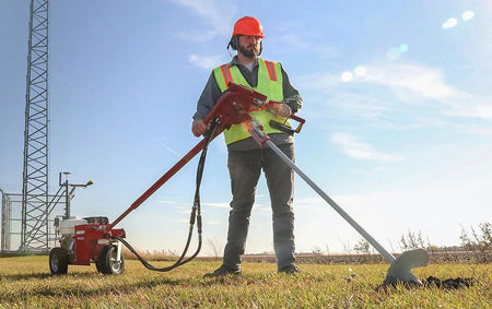 An operational demonstration showing a worker in safety gear using a Little Beaver hydraulic earth drill with a specialized attachment to drive a large ground anchor into a grassy field for structural support.