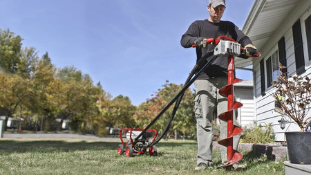 An outdoor application shot of a contractor operating the Little Beaver 5.5 HP post hole digger machine to drill into a residential grass lawn, demonstrating the one-man torque-free safety handlebar setup next to a house.
