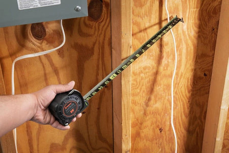 A person's hand holds the Lufkin Shockforce G2 Nite Eye tape measure, extending the black and green blade to measure a wooden stud.