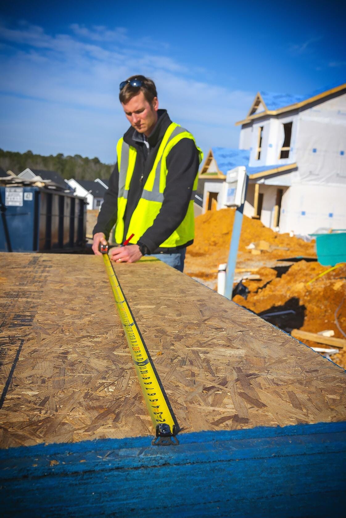A worker in a safety vest uses the Lufkin Shockforce tape measure to measure a large sheet of OSB on a construction site.