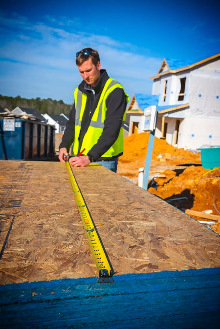 A worker in a safety vest uses the Lufkin Shockforce tape measure to measure a large sheet of OSB on a construction site.