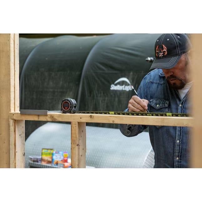 A worker uses the Lufkin Shockforce tape measure, extending the black and yellow blade across a wooden stud to make a precise mark with a pencil during framing.