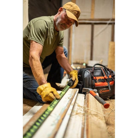 A worker wearing gloves kneels on the floor of a construction site, using the Lufkin Shockforce tape measure to measure a piece of wood trim.