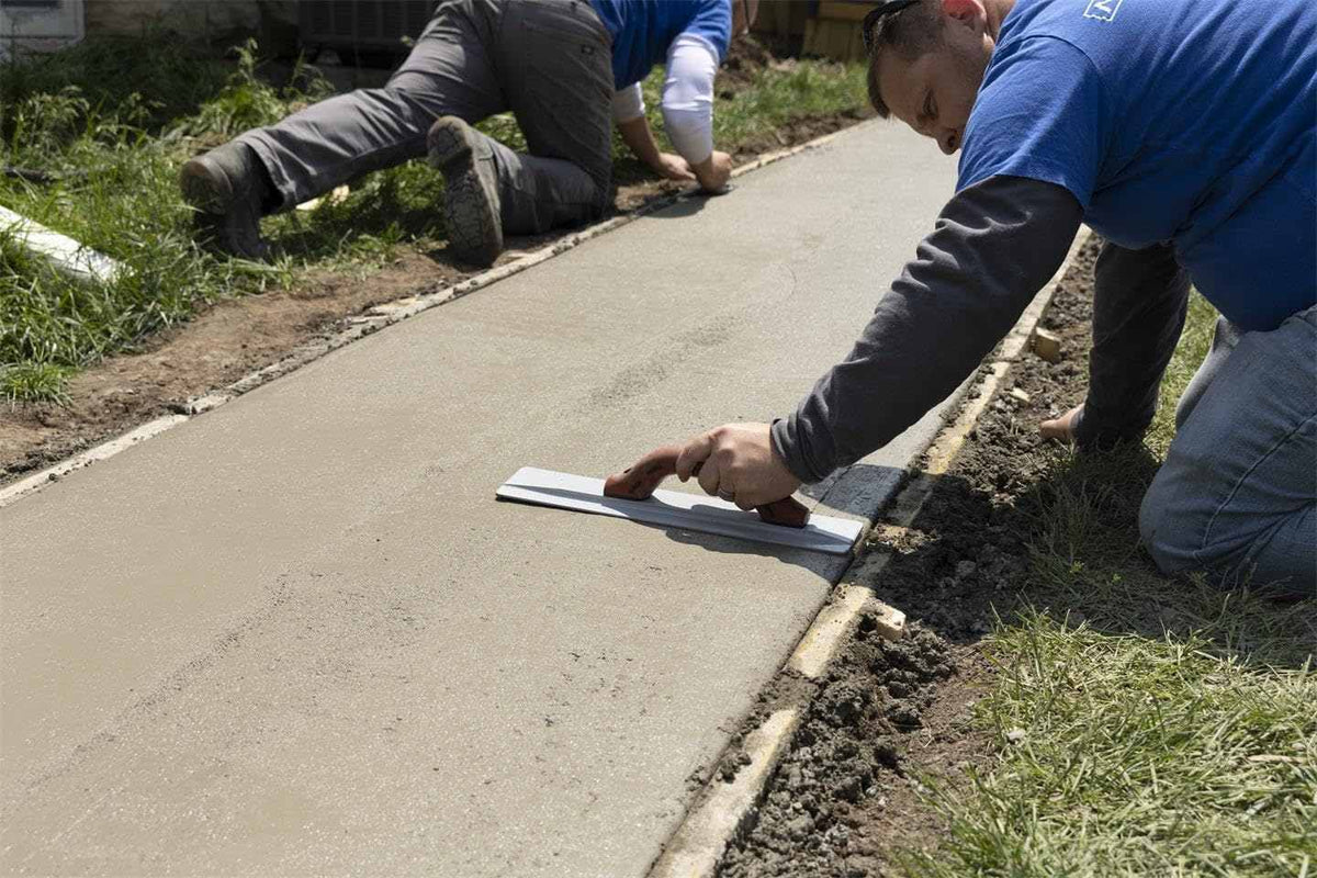 A worker kneels on the grass while using the MARSHALLTOWN magnesium float to smooth the surface of a freshly poured concrete sidewalk.