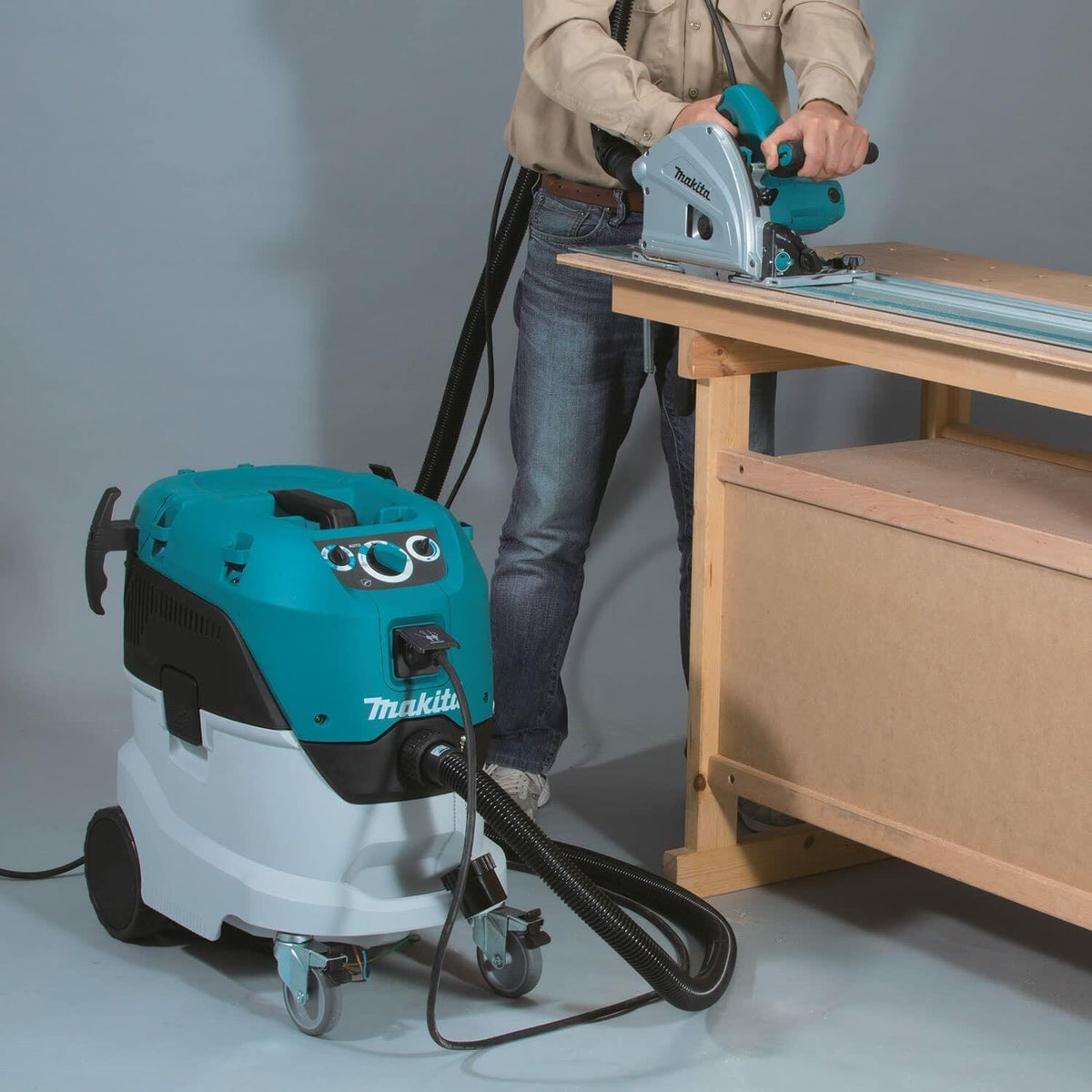 Makita VC4210L dust extractor connected to a track saw during wood cutting on a workbench, demonstrating dust collection in a woodworking setup.