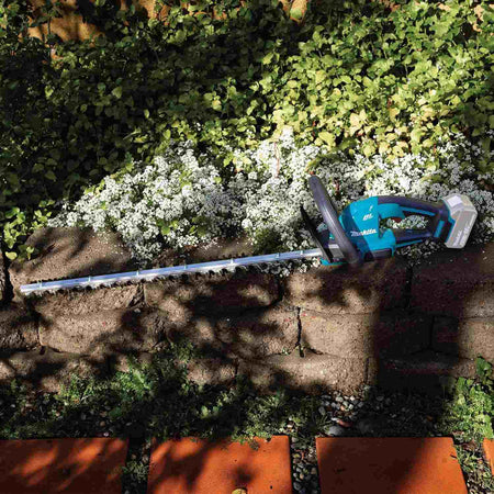 Makita XHU09Z 18V LXT brushless hedge trimmer resting on a stone retaining wall in a garden, showcasing its cordless portability and 24-inch blade in an outdoor landscaping environment.