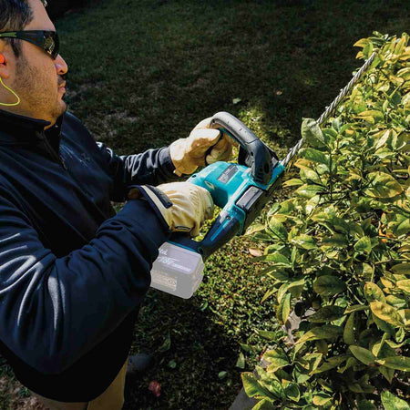 Contractor using the Makita 18V cordless hedge trimmer with an attached LXT lithium-ion battery to perform precision pruning on a dense green bush.