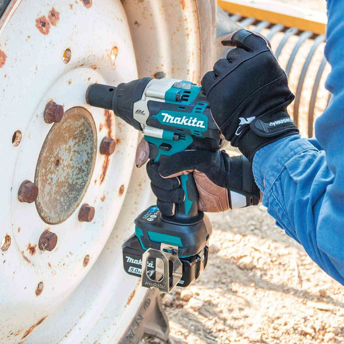 A person in gloves uses the Makita XWT18T impact wrench with a deep socket to remove a large, rusty bolt from the wheel of a tractor or heavy machinery.