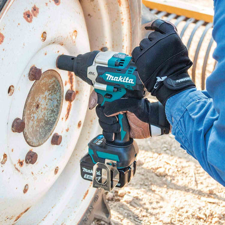 A person in gloves uses the Makita XWT18T impact wrench with a deep socket to remove a large, rusty bolt from the wheel of a tractor or heavy machinery.