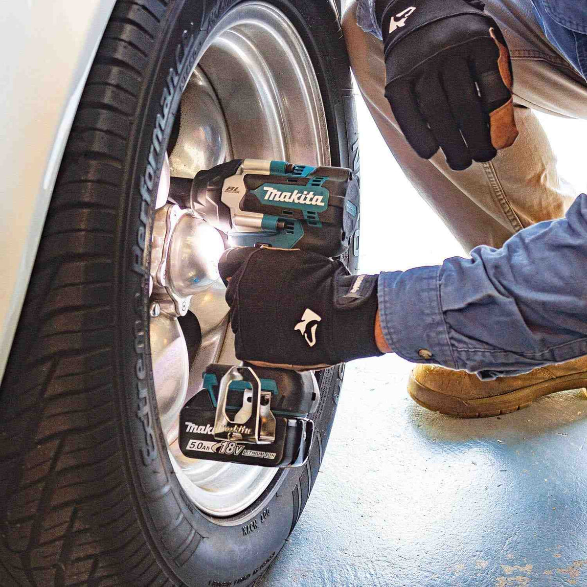 A person in gloves uses the Makita XWT18T 1/2-inch impact wrench to remove the lug nuts from a car's wheel, with the tool's LED light illuminating the work area.