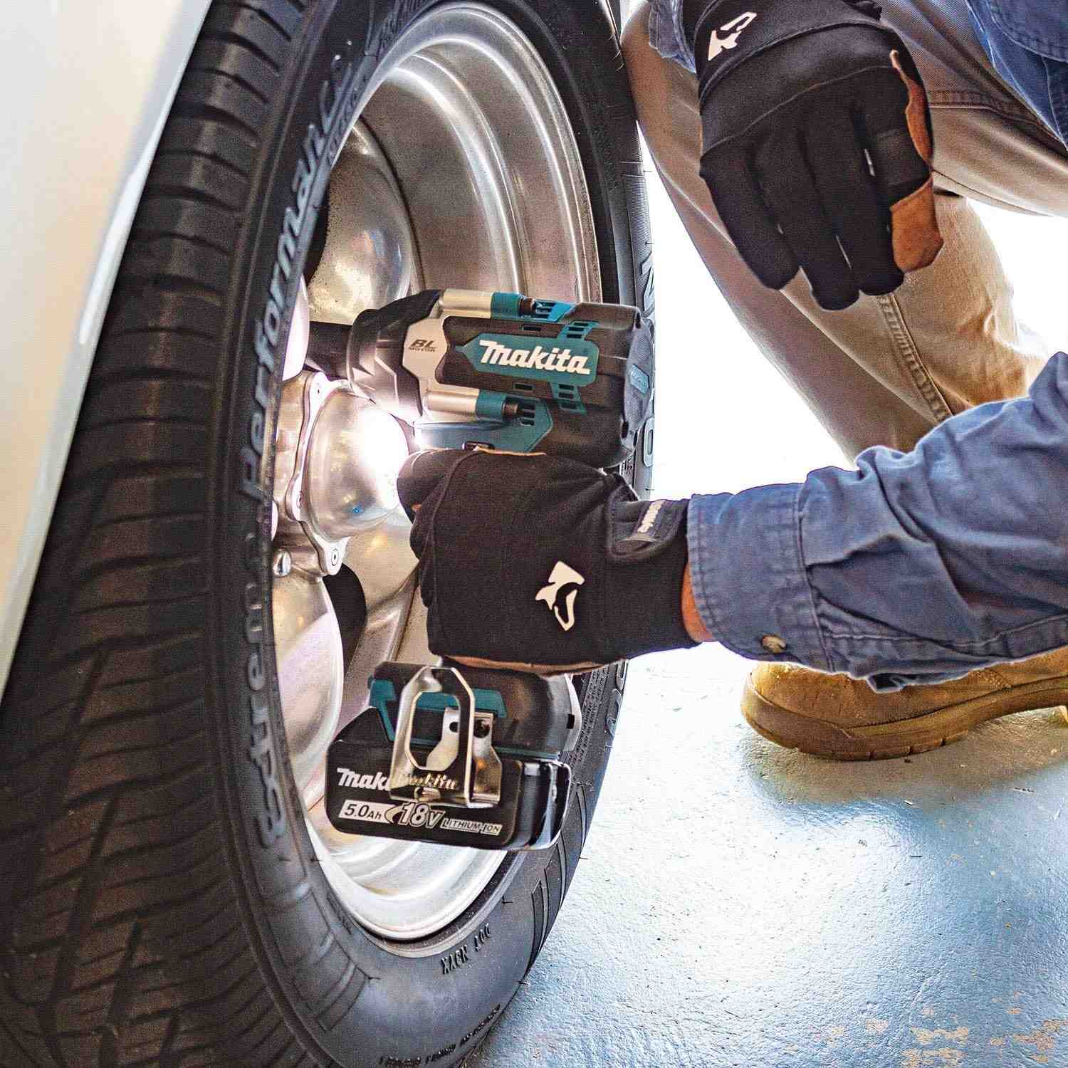 A person in gloves uses the Makita XWT18T 1/2-inch impact wrench to remove the lug nuts from a car's wheel, with the tool's LED light illuminating the work area.
