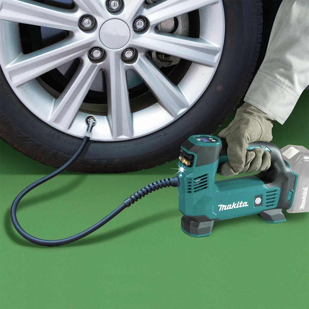 Makita portable inflator connected to a car tire valve; person wearing gloves operates the tool on a green surface with silver alloy wheel in view.