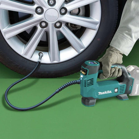 Makita portable inflator connected to a car tire valve; person wearing gloves operates the tool on a green surface with silver alloy wheel in view.