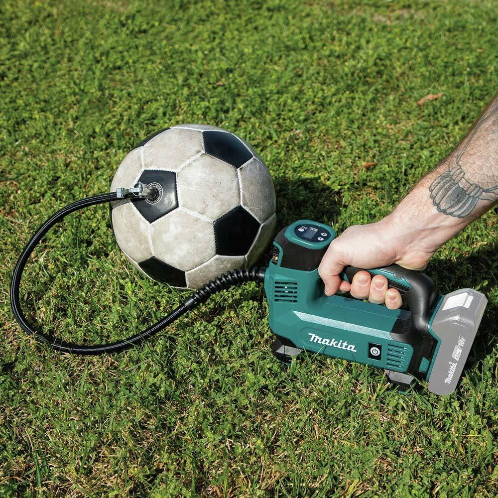 Makita cordless inflator in use outdoors inflating a soccer ball; hose attached to valve with battery pack visible and grassy surface beneath.