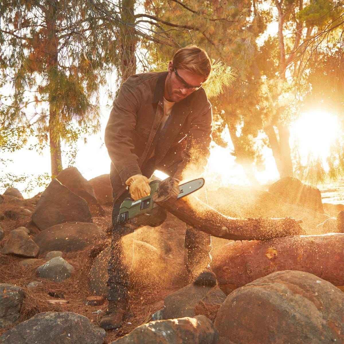 Action shot of a worker using the Makita 18V LXT Outdoor Adventure chain saw to prune a fallen tree branch at sunset, demonstrating gas-equivalent performance and low noise.