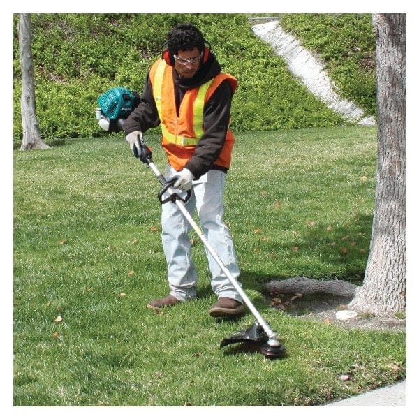 Person wearing safety gear using a string trimmer to cut grass around a tree in a landscaped area. High-visibility vest, gloves, and ear protection worn. Sidewalk and trees visible in background.