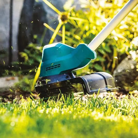 Close-up of string trimmer actively cutting grass near a stone border. Green and black casing visible. Grass clippings flying through air in outdoor garden setting
