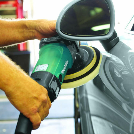 Close-up action shot of a person's hands guiding the Makita 7-inch polisher as it buffs the area around the side mirror of a silver car.