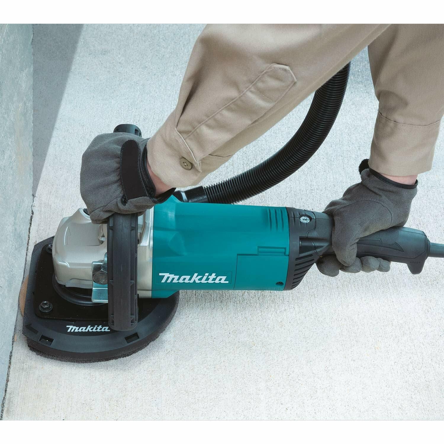 Person operating Makita concrete planer near wall; tool connected to vacuum hose with dust shroud, user wearing gloves and long-sleeved shirt for safety.