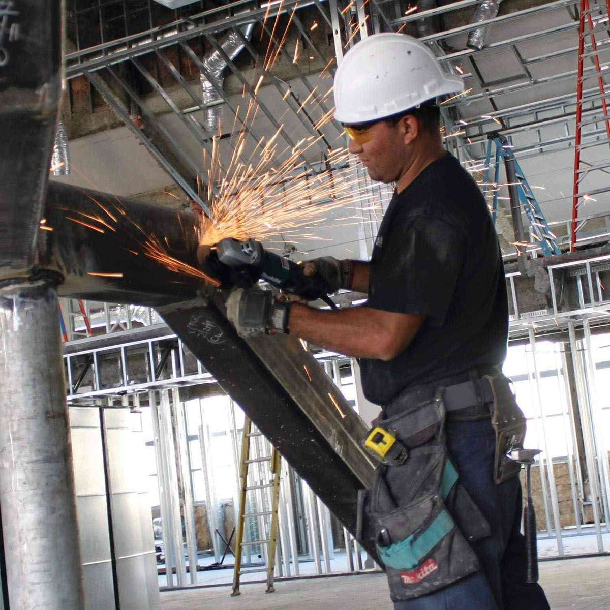 A professional construction worker in a hard hat and safety gear using the Makita 9565PCV angle grinder to perform overhead metal grinding on a jobsite steel structure.