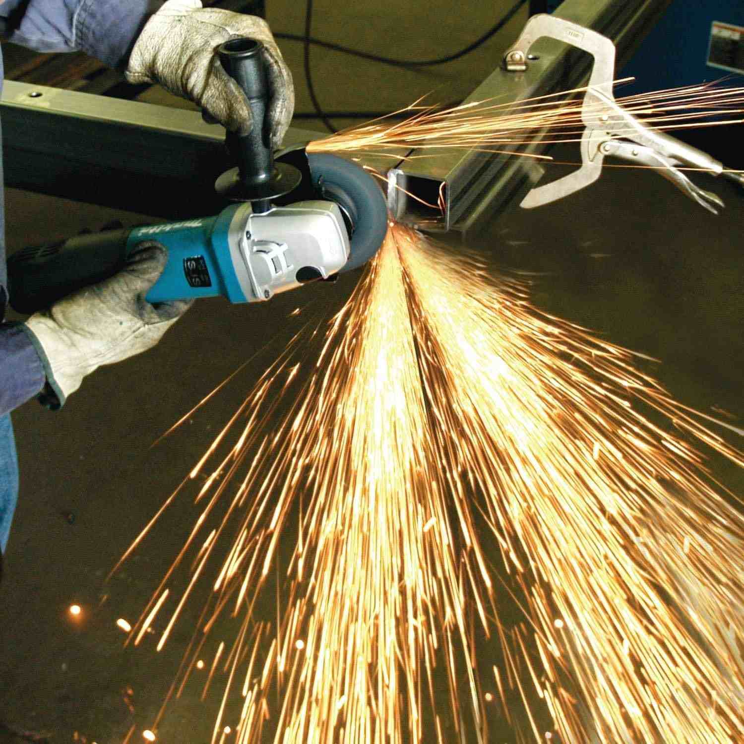 Action shot showing the Makita 5-inch angle grinder being used with a side handle for stability while cutting through metal framing, highlighting the tool's precision and high output.