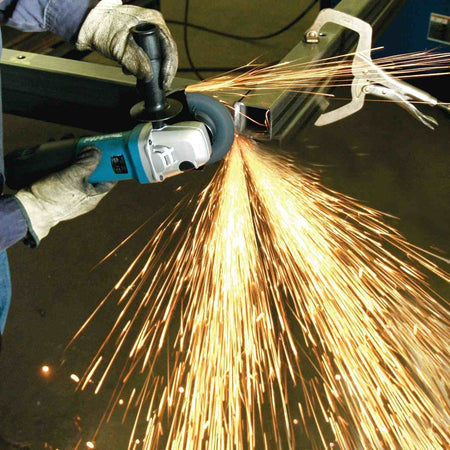 Action shot showing the Makita 5-inch angle grinder being used with a side handle for stability while cutting through metal framing, highlighting the tool's precision and high output.