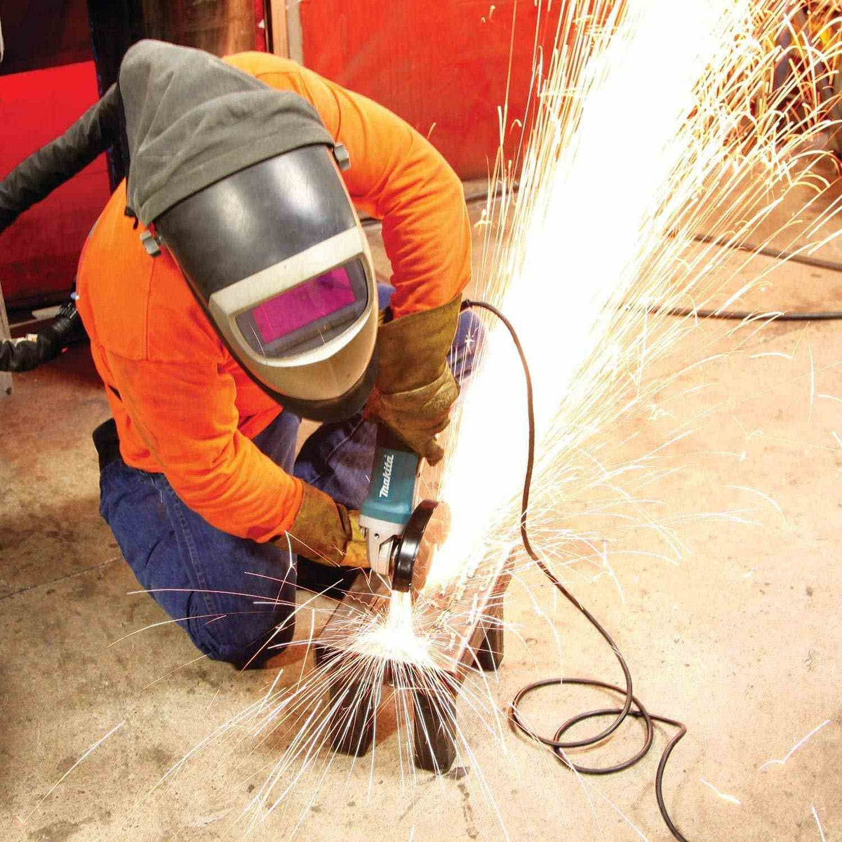 A professional welder wearing a protective hood using the Makita 13 Amp corded angle grinder to finish a metal workpiece, showcasing the tool's reliability in extreme industrial environments.