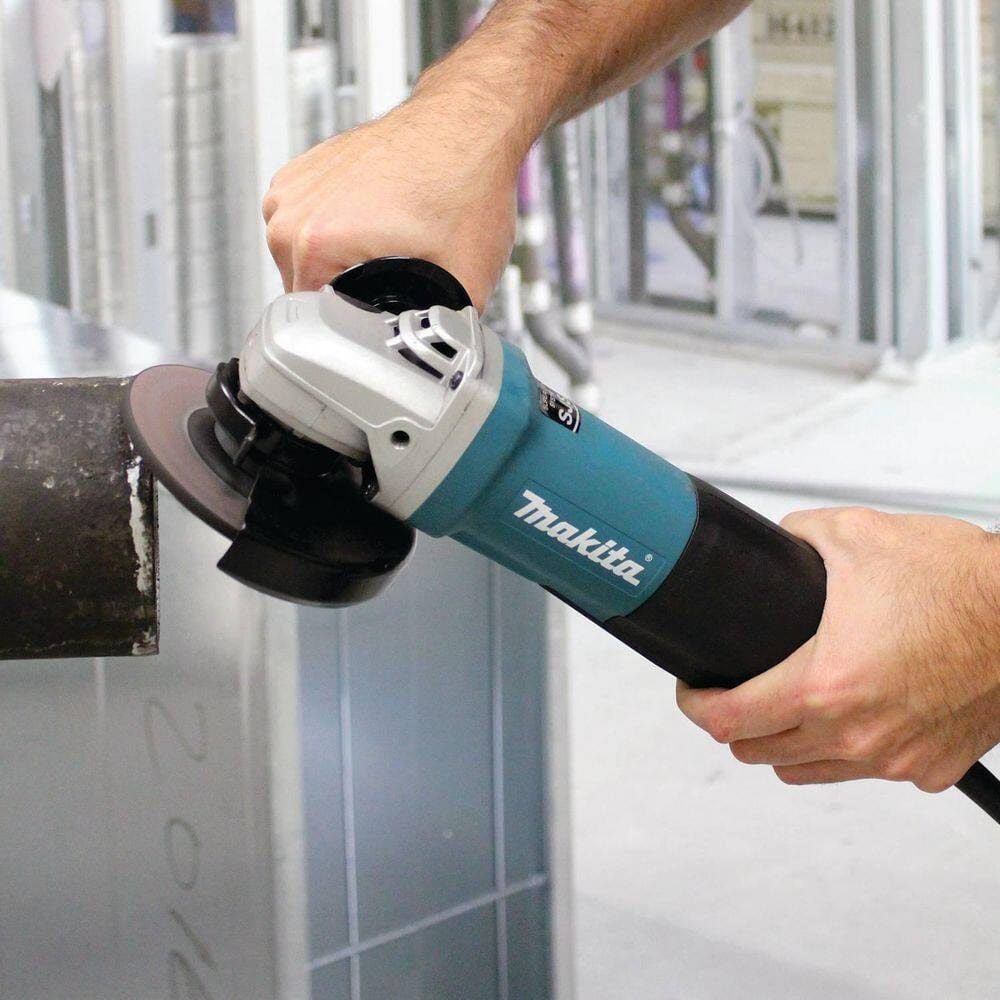 Action photograph showing a professional tradesperson using the Makita 5-inch angle grinder to clean and prep a heavy-duty metal pipe for welding.