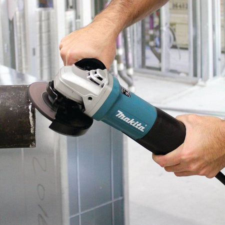 Action photograph showing a professional tradesperson using the Makita 5-inch angle grinder to clean and prep a heavy-duty metal pipe for welding.