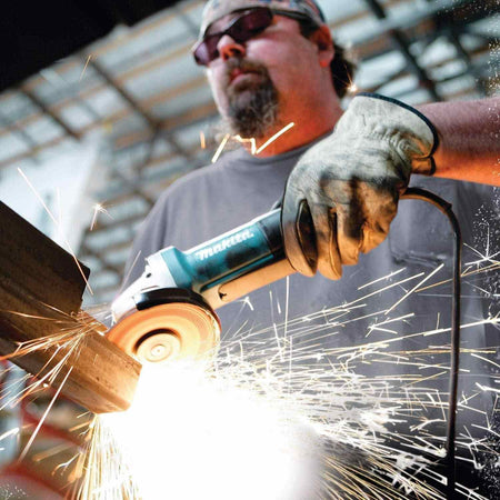 Action photograph of a professional using the Makita 13 Amp high-power angle grinder to grind a steel beam, showing a high volume of sparks and demonstrating industrial performance and control.