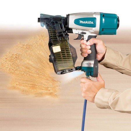 Action shot demonstrating the powerful built-in air duster on the Makita AN613 pneumatic siding coil nail gun actively blowing debris away from a wood framing workspace.