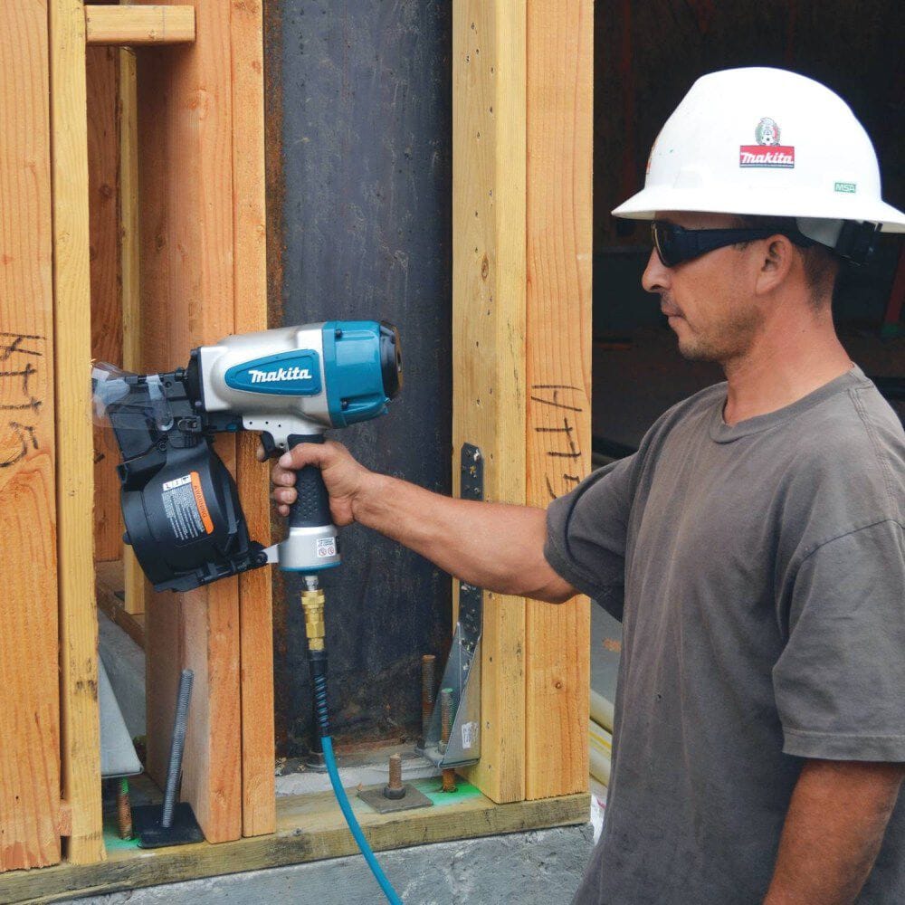 A professional contractor wearing a hard hat and safety glasses holding the Makita AN902 pneumatic coil framing nailer while standing next to wooden structural framing.