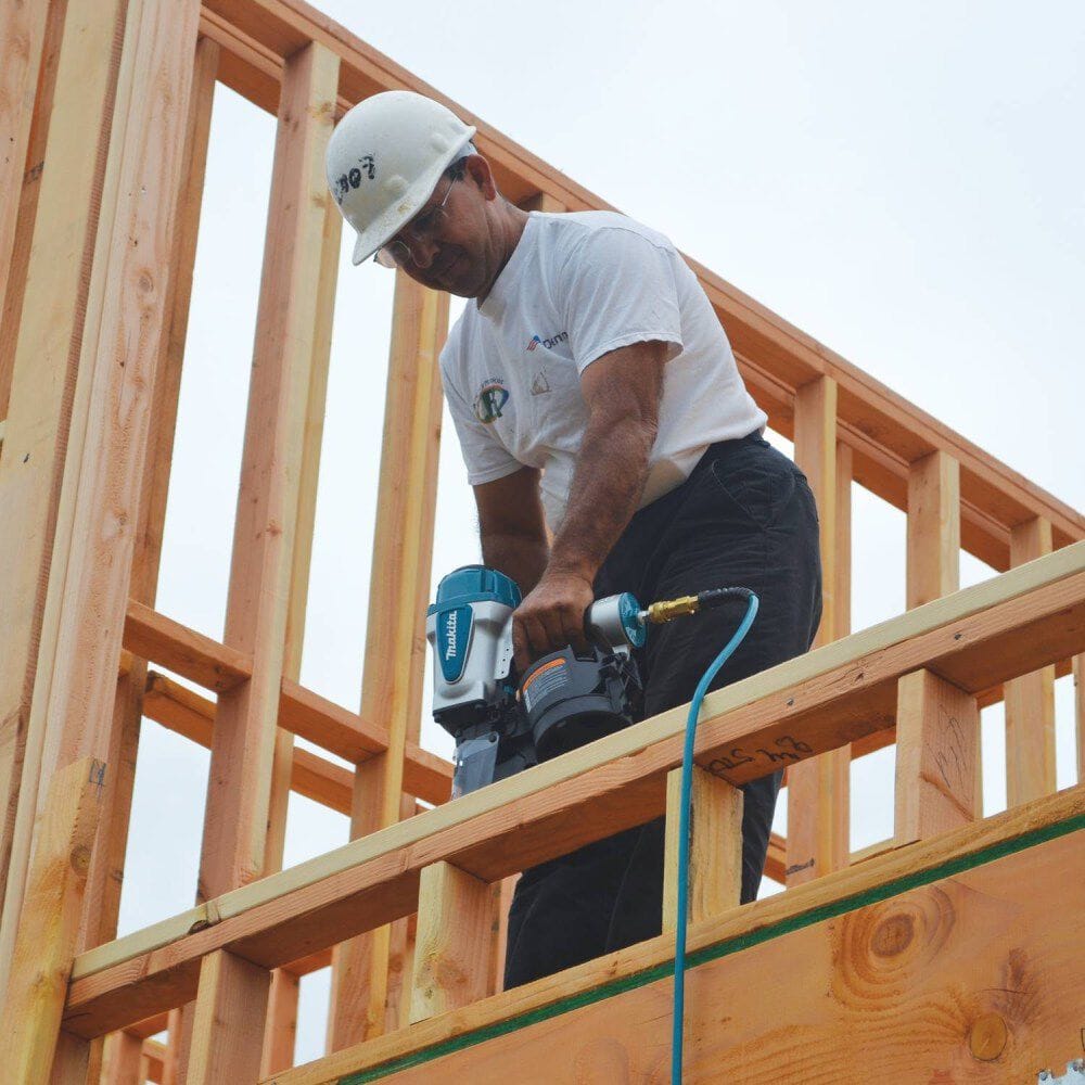 A woodworker operating the Makita AN902 coil framing nailer to drive fasteners into an upper wooden beam during a home construction project.