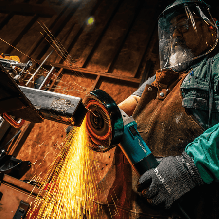 Side angle of a tradesperson cutting thick steel stock with the Makita 9566CVR angle grinder in a workshop.