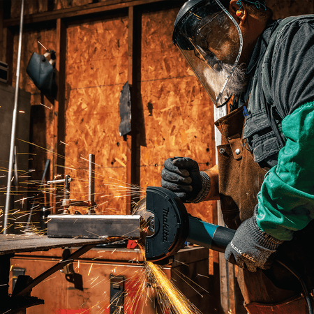 Metal fabricator wearing a protective face shield operating the Makita cut-off grinder on a structural steel workpiece.
