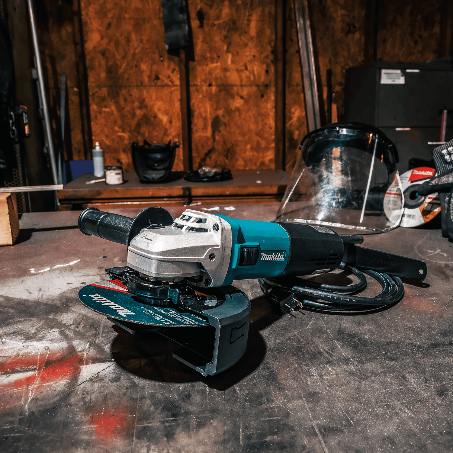 Makita 9566CVR angle grinder resting on a workshop welding table alongside safety gear and a cut-off wheel.