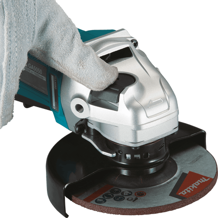 Gloved thumb depressing the large spindle lock button on the cast aluminum gear head of the Makita GA4553R angle grinder.