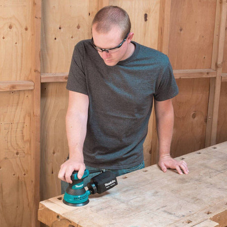 Man using a Makita sander on a wooden surface with a wooden wall background