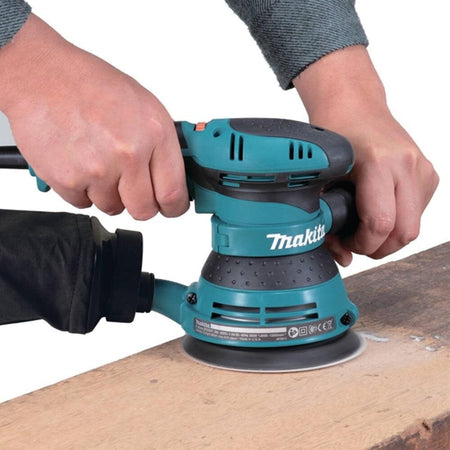 Action shot of a professional woodworker using the Makita 3 AMP random orbit sander with both hands on a piece of dimensional lumber, demonstrating the superior stability provided by the dual-handle design.