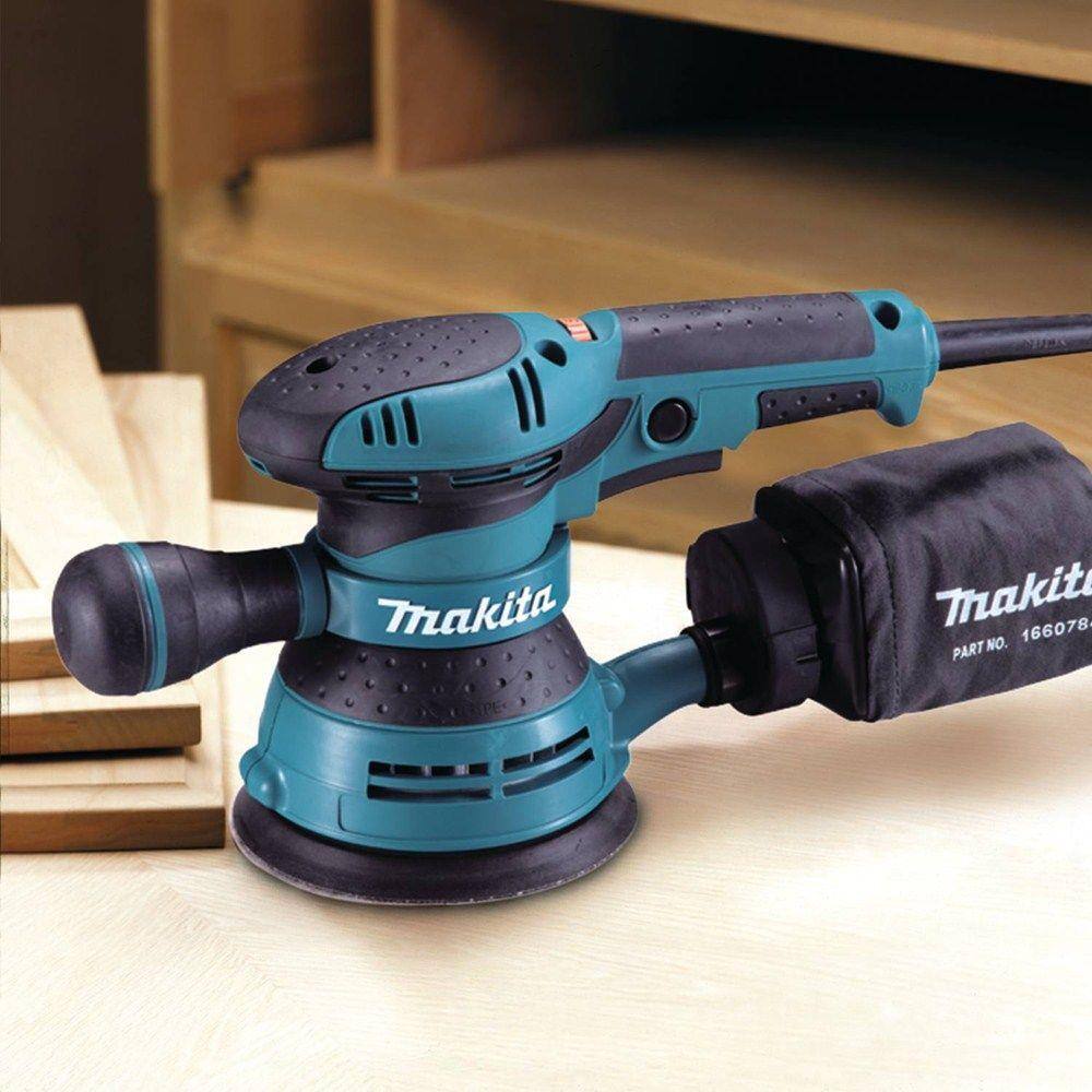 Side profile view of the Makita BO5041 variable speed sander resting on a wooden workpiece in a workshop setting, emphasizing its ergonomic build and readiness for high-precision finish work.