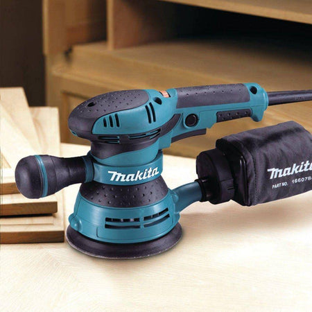 Side profile view of the Makita BO5041 variable speed sander resting on a wooden workpiece in a workshop setting, emphasizing its ergonomic build and readiness for high-precision finish work.