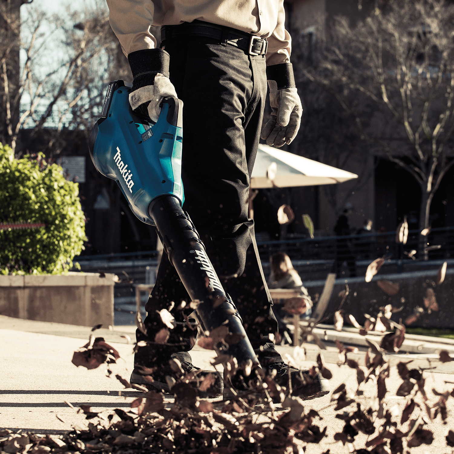 Groundskeeper using the quiet 61 dB(A) Makita XBU02PT 36V cordless blower to clear leaves and debris off a concrete commercial walkway.