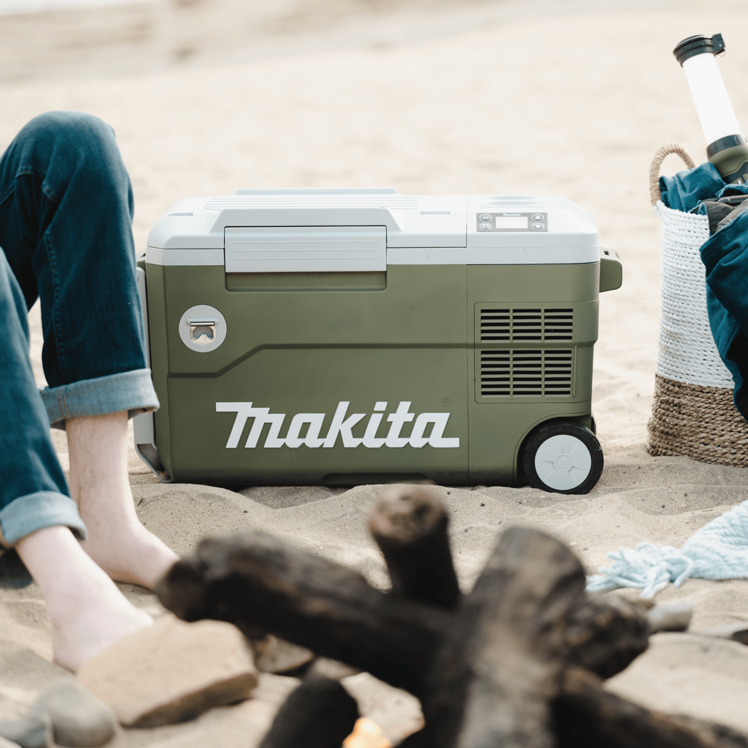 Makita ADCW180Z Outdoor Adventure 21-quart cooler and warmer resting on a sandy beach near a campfire setup.