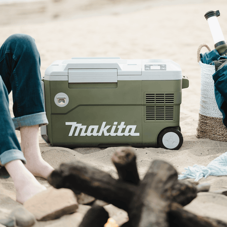 Makita ADCW180Z Outdoor Adventure 21-quart cooler and warmer resting on a sandy beach near a campfire setup.