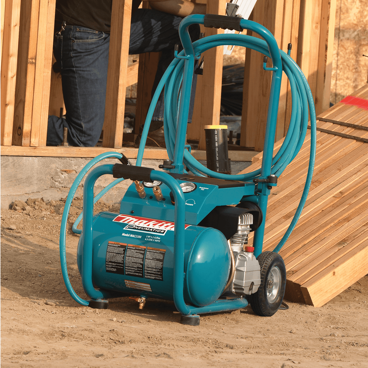 Makita MAC5200 wheeled air compressor with wrapped 50-foot air hose parked near lumber on a framing construction site.