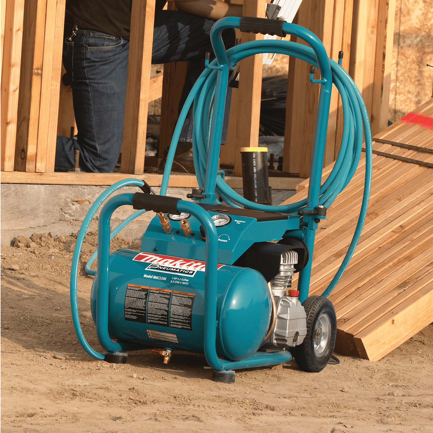 Makita MAC5200 wheeled air compressor with wrapped 50-foot air hose parked near lumber on a framing construction site.