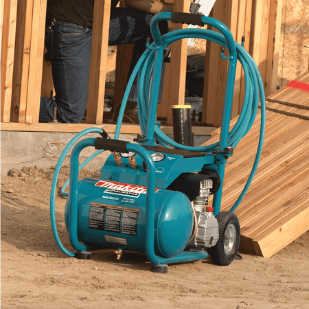 Makita MAC5200 wheeled air compressor with wrapped 50-foot air hose parked near lumber on a framing construction site.